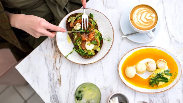 Woman eating her tasty brunch hipster cafe top view marble table salmon avocado toast coffee sweet tasty cheesecakes enjoying her breakfast