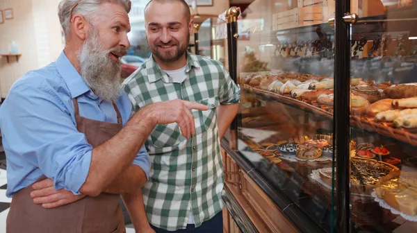 Young man talking senior baker while shopping pastry local bakery